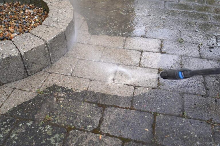Technician cleaning a concrete walkway beside a bright residential entry with palms and outdoor walls.